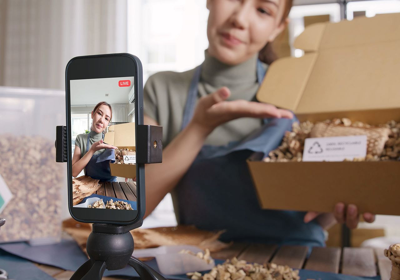 A small business owner in an apron shows a product box with recyclable packaging during a live stream filmed on a smartphone tripod.