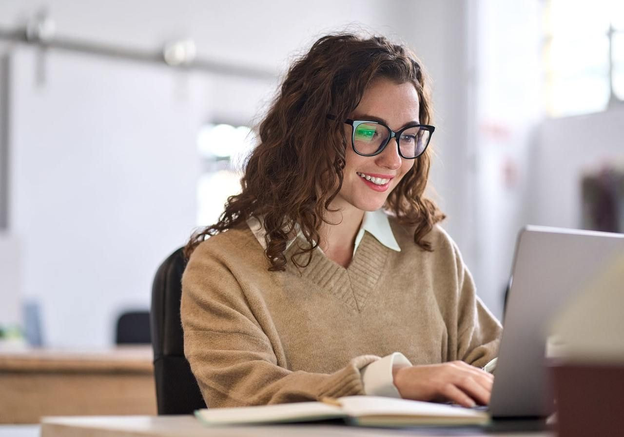 A young woman in glasses is looking at a laptop screen and smiling.