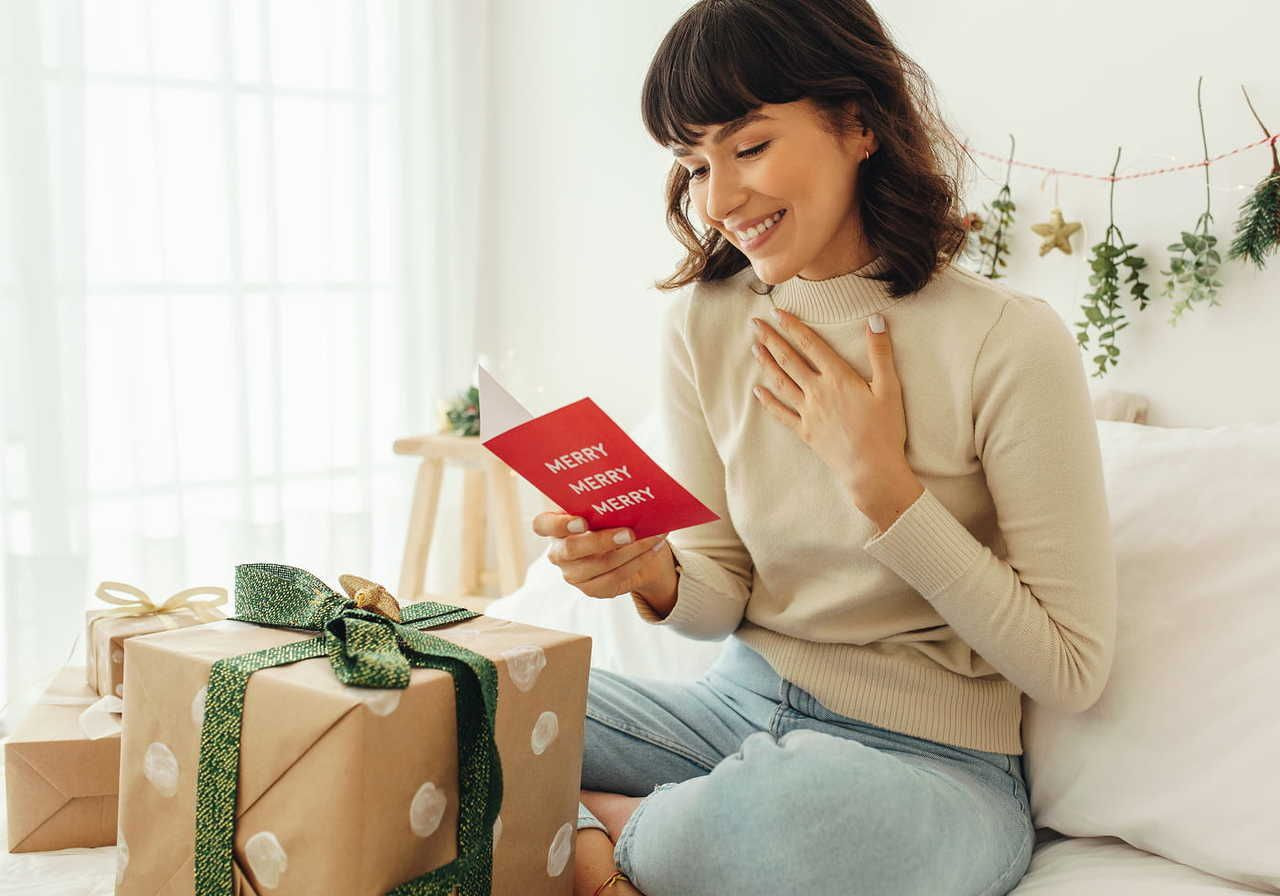 A woman in a beige sweater and jeans smiles while reading a red card saying "Merry Merry Merry." She is sitting on a bed with wrapped gifts nearby.