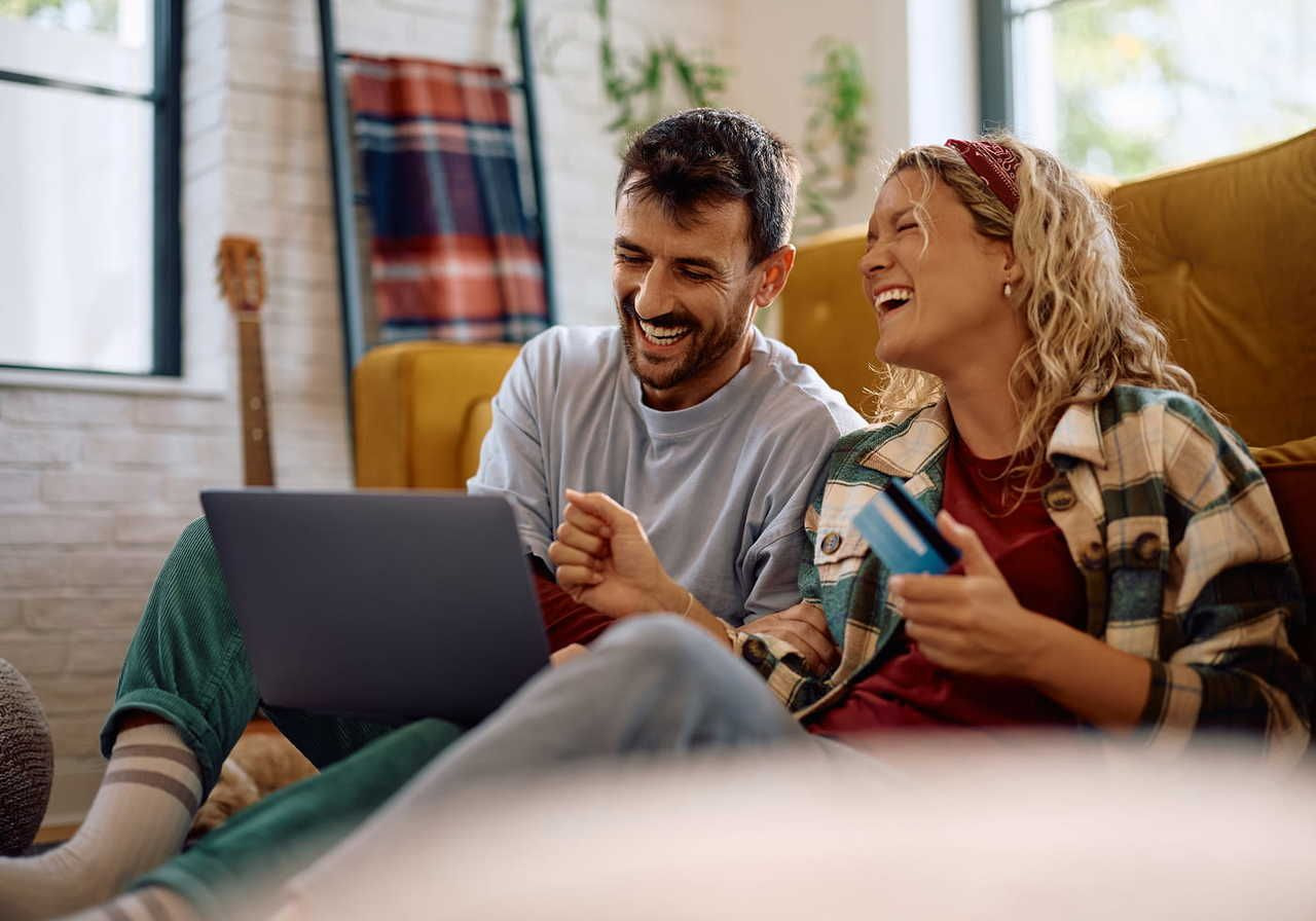 Two people sitting on the floor, laughing together while using a laptop.