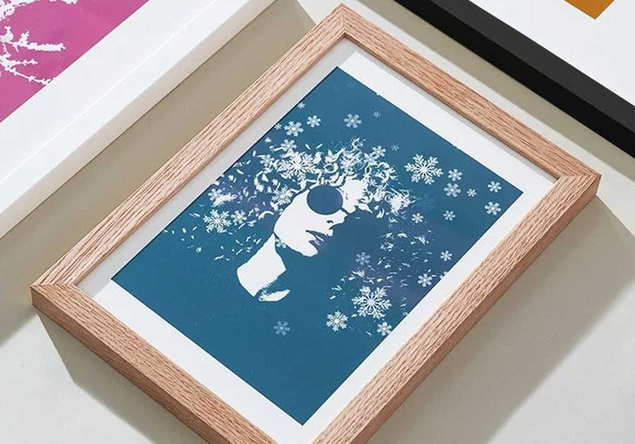 Framed artwork featuring a stylized portrait with curly hair and sunglasses, set against a blue background with snowflake patterns.