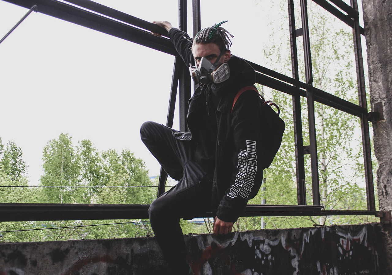 A person with green braided hair wearing a gas mask and a black hoodie sits on a ledge in an urban environment with greenery in the background.