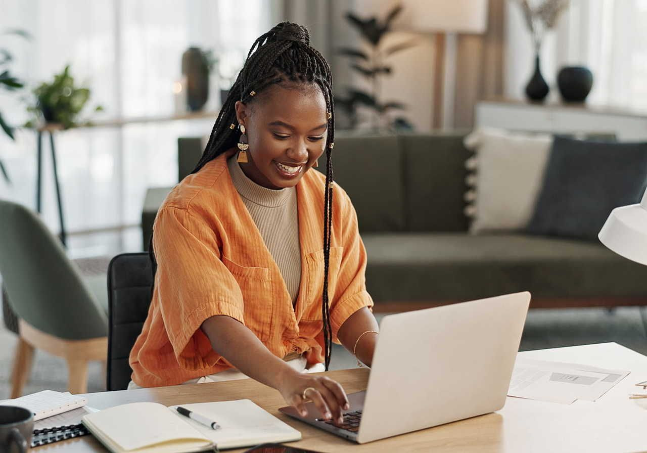 woman with braided hair sitting at a desk, working on a laptop, smiling and looking engaged. She is wearing an orange shirt.