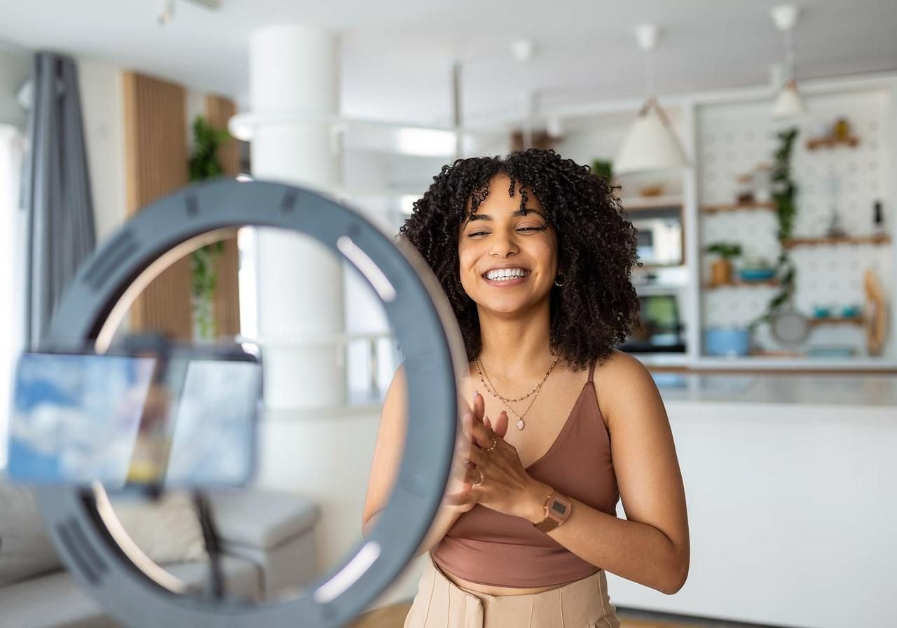 Smiling woman with curly hair stands in a modern kitchen, clapping, captured by a smartphone on a tripod with ring light.