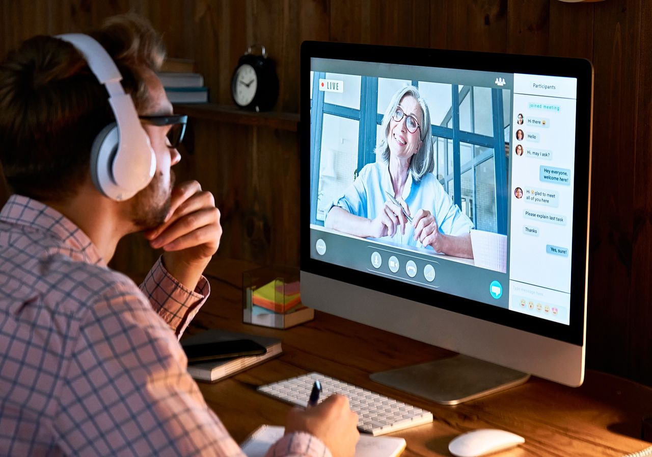 A man with headphones participates in a video call on a computer, showing a woman in glasses. 