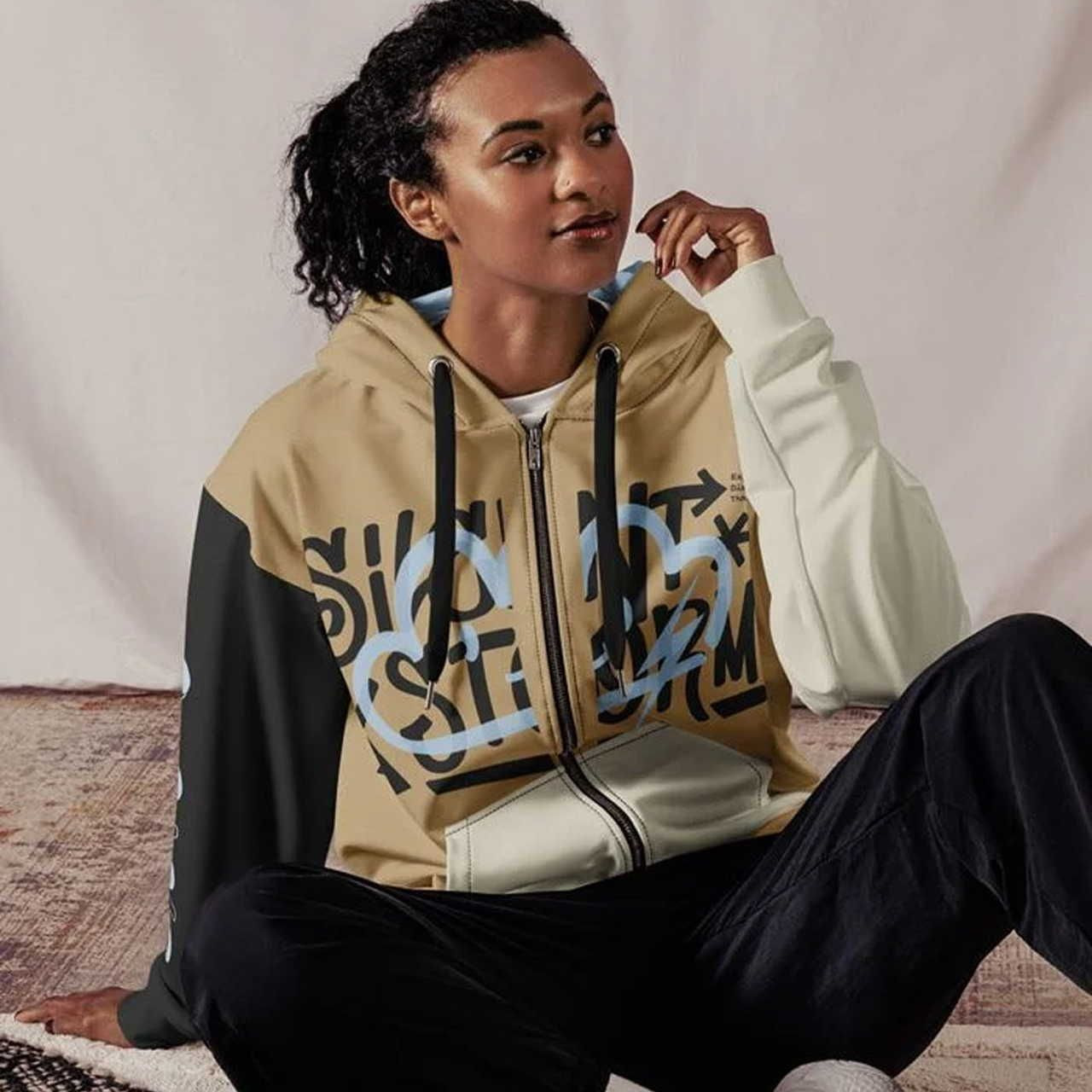 A woman with curly hair sits casually on the floor wearing a multicolored hoodie with abstract lettering.