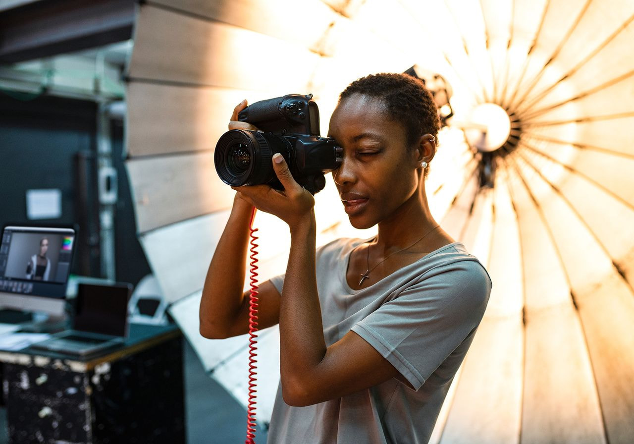 A focused photographer with a digital camera stands in a studio, using a lighting umbrella in the background.