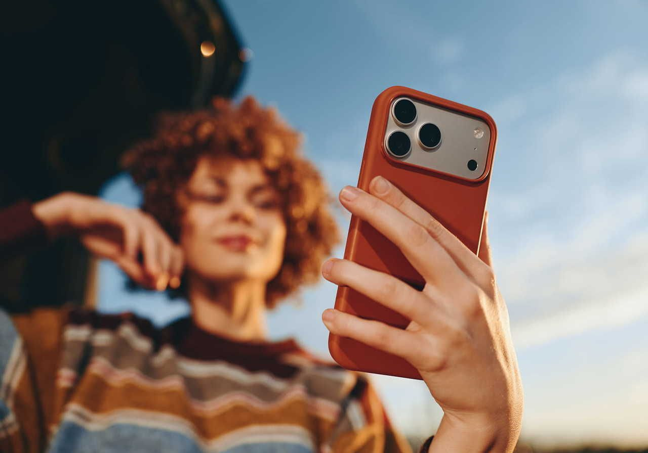 A woman with curly hair is holding a smartphone while looking at it, with a blurred landscape in the background.