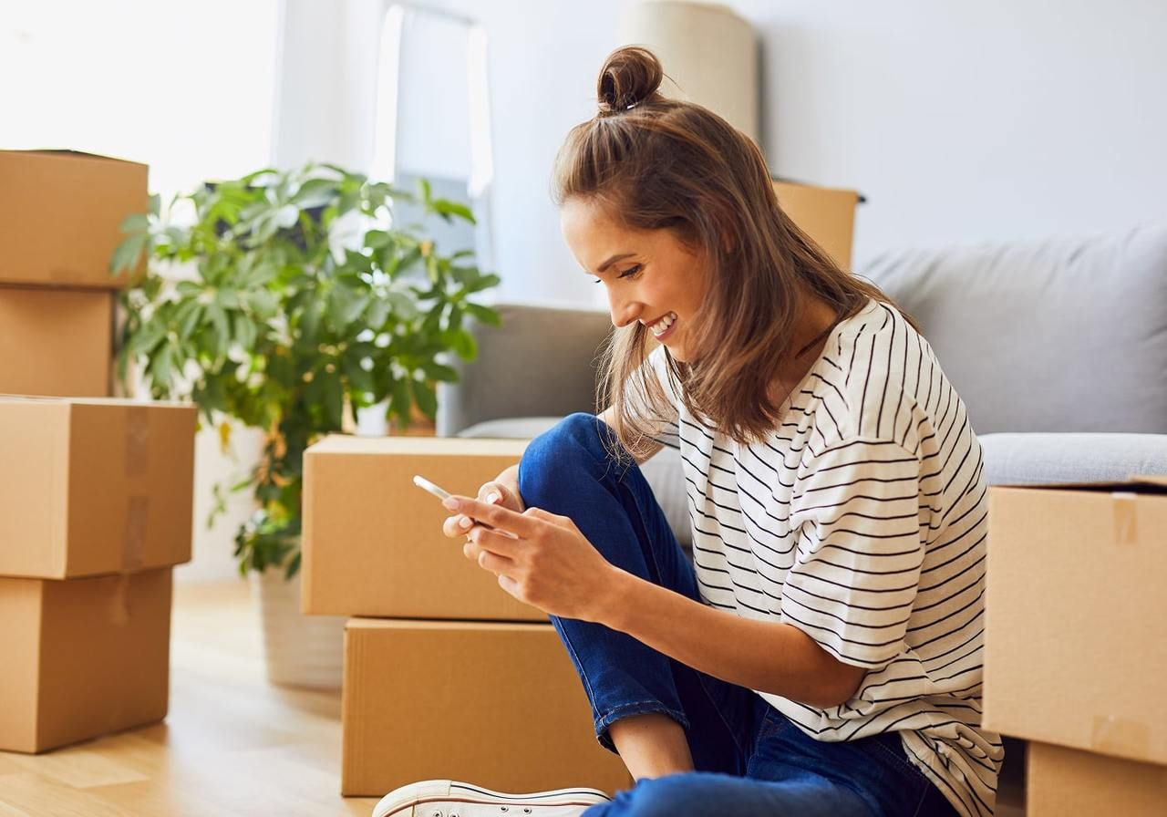 A woman sits on the floor among moving boxes, smiling as she looks at her phone, learning about TikTok dropshipping.