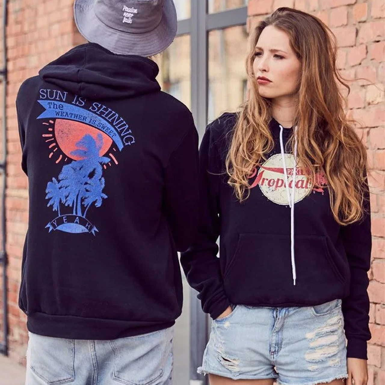 Two people in casual attire stand against a brick wall. One wears a black hoodie with a tropical graphic, while the other has a sun graphic.