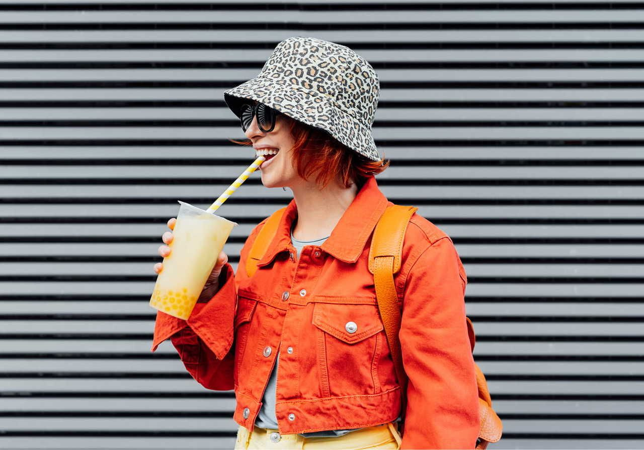 A woman in an orange jacket and leopard print hat smiles while sipping bubble tea through a straw.