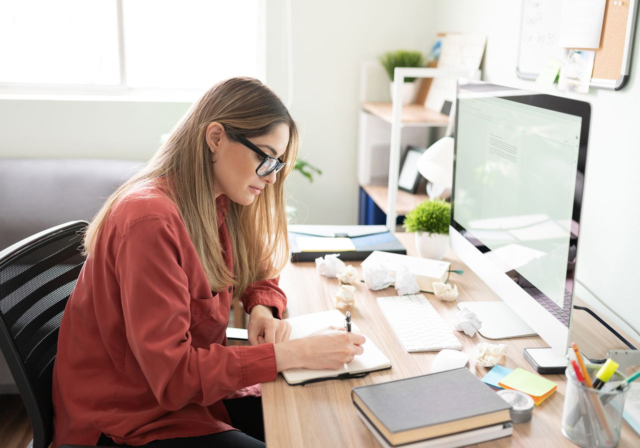 A woman sits at a desk with a computer and a notebook, focused on her work