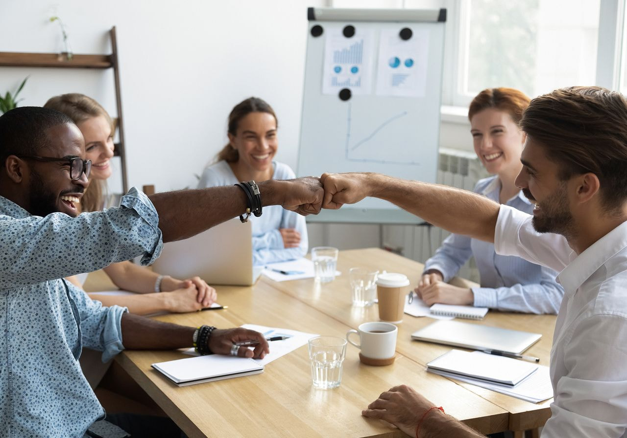Two smiling coworkers fist-bump across a conference table as teammates celebrate in front of a whiteboard with growth charts.