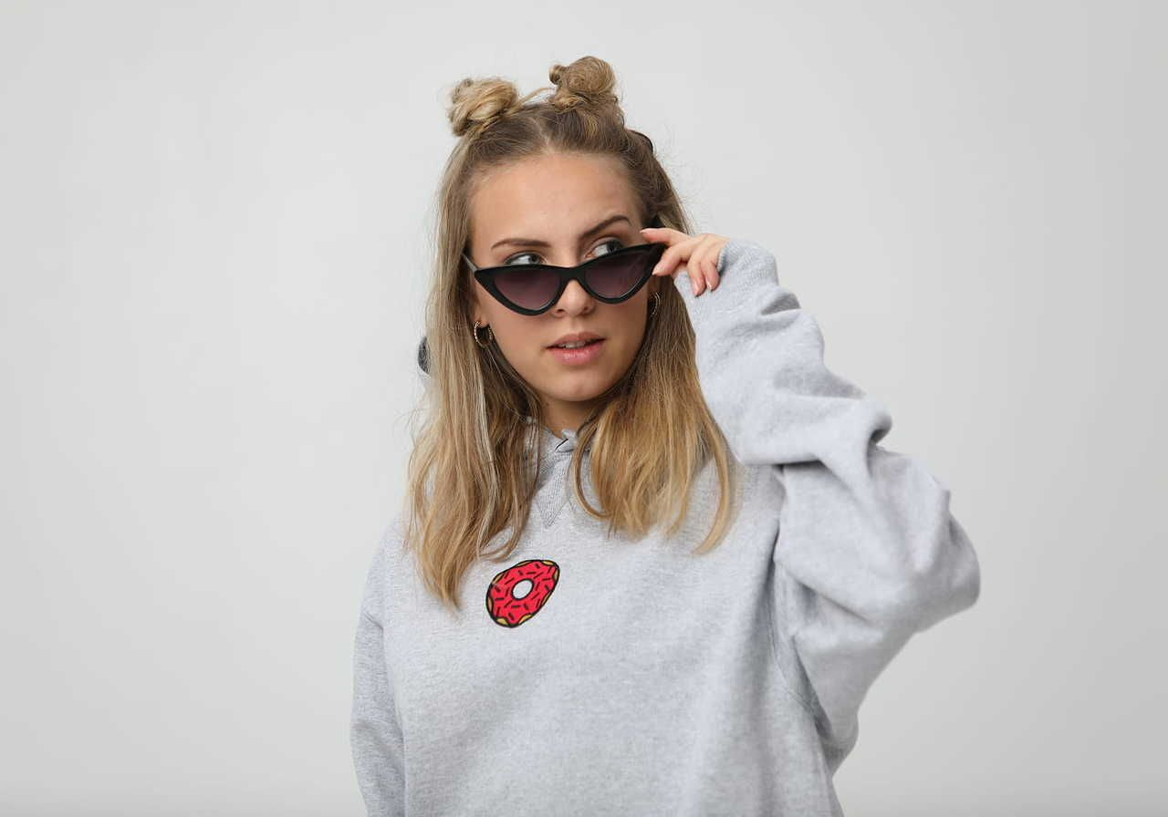 A young woman with two buns in her hair is wearing a gray hoodie with an embroidered donut design, adjusting her dark sunglasses.