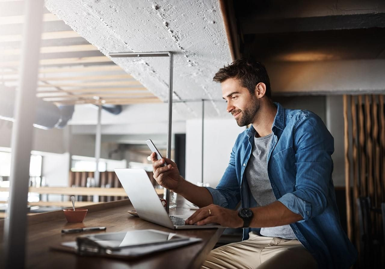 A man in a casual blue shirt smiles while working at a laptop in a modern cafe, holding a smartphone.