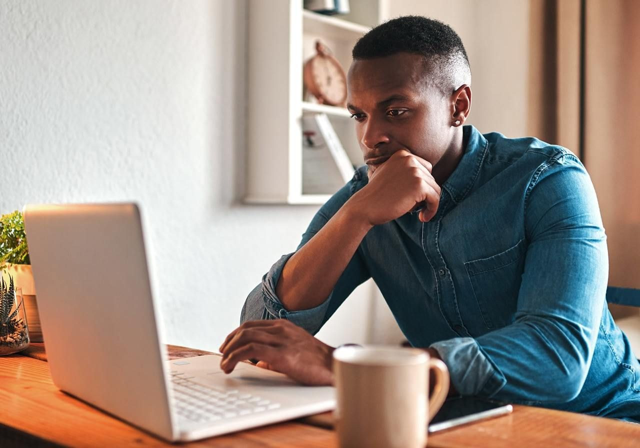 A man in a denim shirt sits at a wooden desk, focused on a laptop, searching for how to make money on Canva.