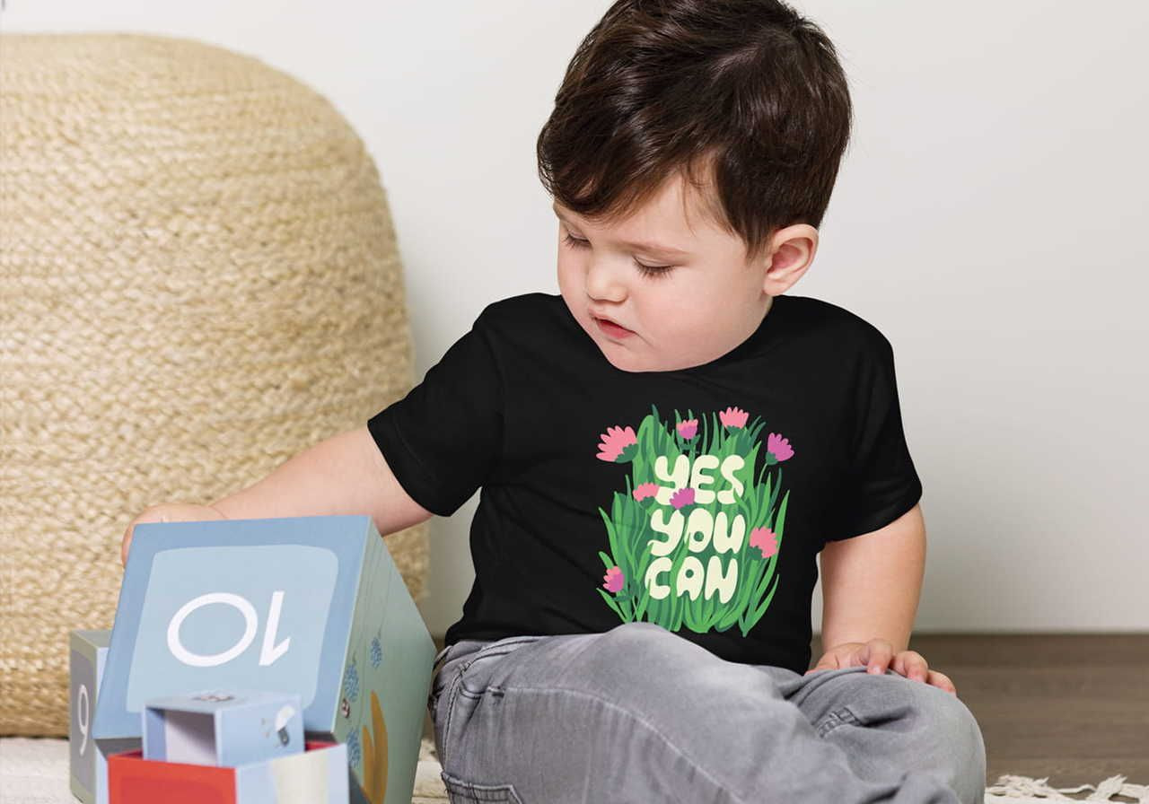 A toddler wearing a black shirt with "Yes You Can" surrounded by flowers, looks at colorful blocks.