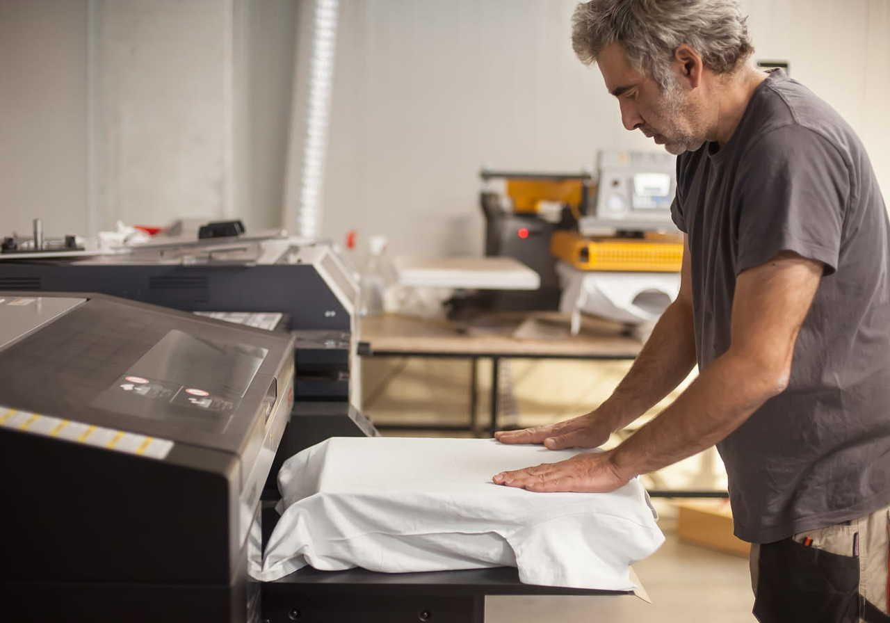 A man is preparing to print on a white t-shirt using a large DTG printer in a workshop.