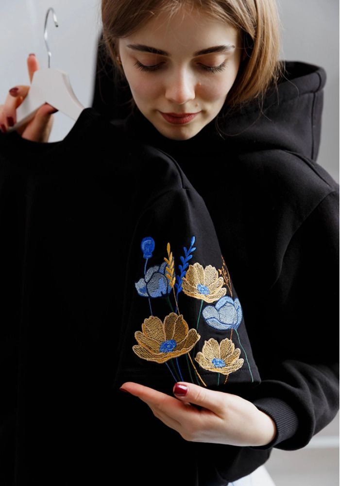 A woman holds a black sweatshirt adorned with colorful embroidered flowers, showcasing a blend of style and nature.