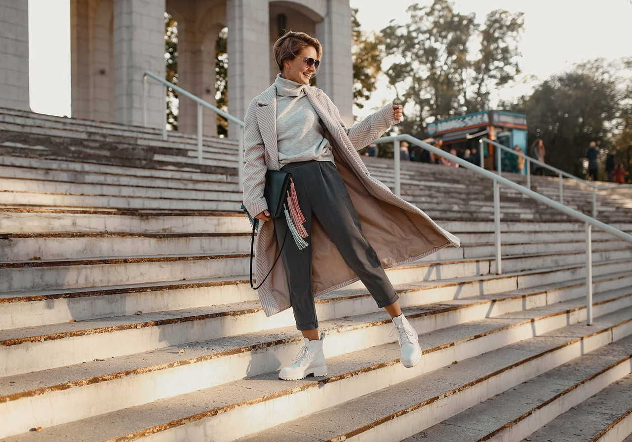 A woman in a long coat and sunglasses poses confidently on wide stone steps in sunlight. The background shows trees and a distant structure.