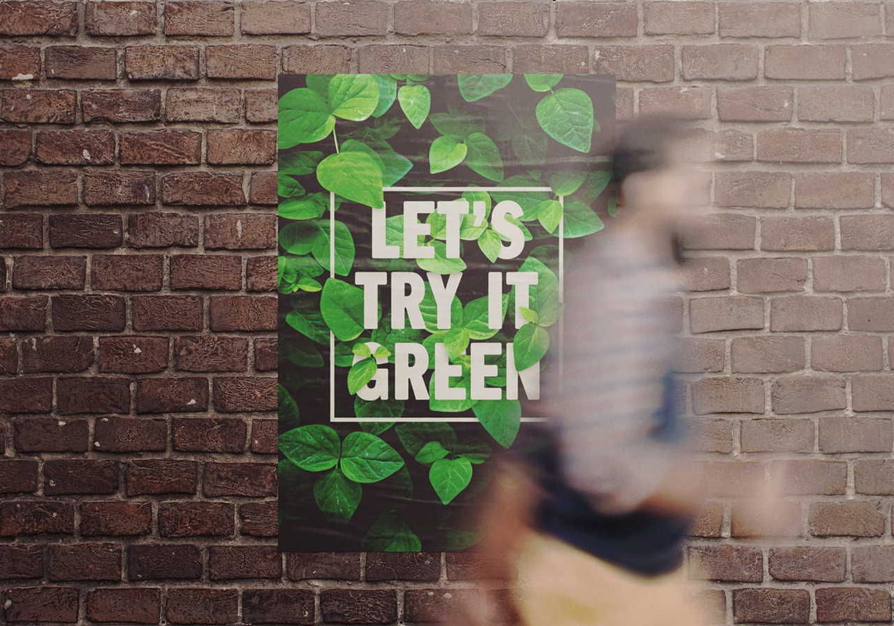 Poster on a brick wall with green leaves and white text saying "Let’s Try It Green." A blurred person walks by.