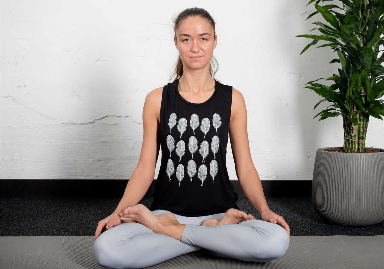 A woman sitting cross-legged in a yoga pose, wearing a black tank top with a graphic design and light gray leggings.