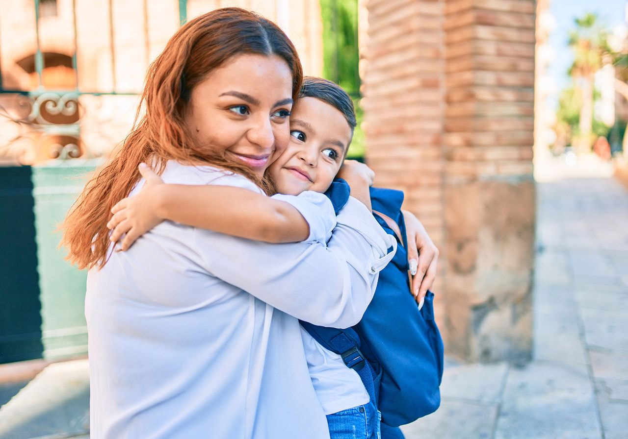 A woman and a child embrace warmly outside, surrounded by greenery and sunlight.