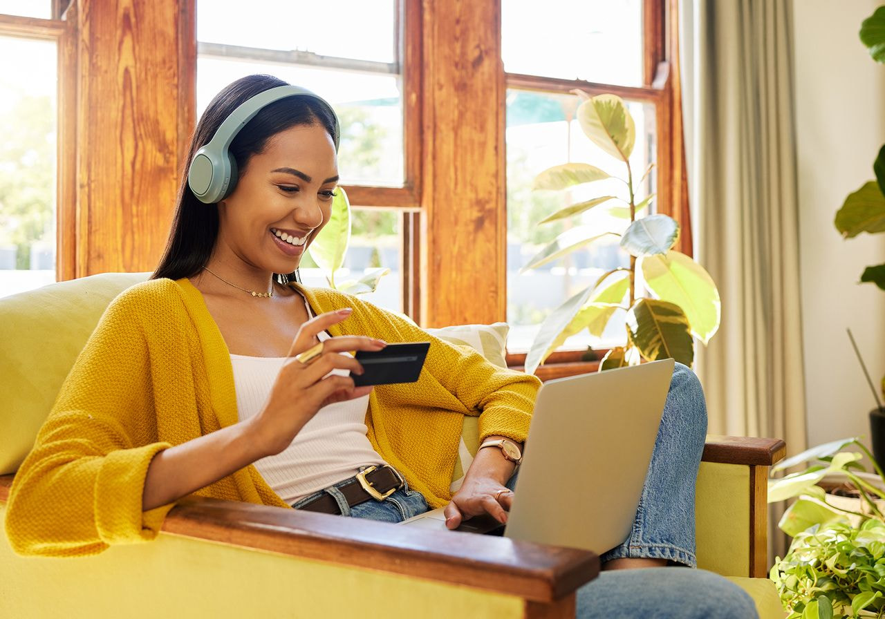 A smiling woman wearing headphones holds a credit card while shopping online on a laptop from a yellow armchair.