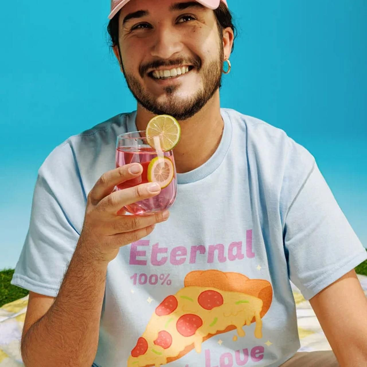 A smiling man in a pink cap and sky-blue t-shirt with pizza graphic, holding a pink drink garnished with lemon.