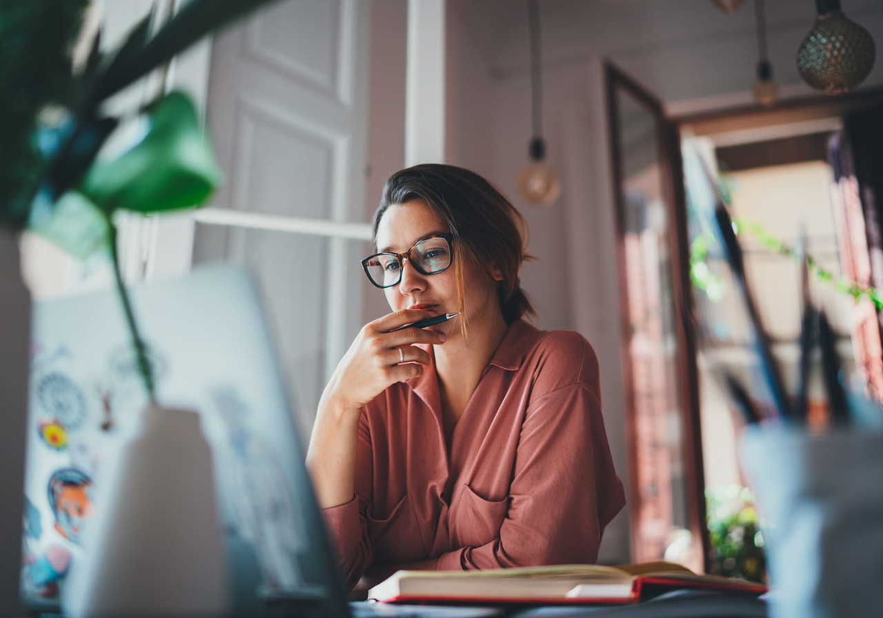 A woman with glasses sitting at a desk, looking thoughtfully at her laptop while holding a pen.