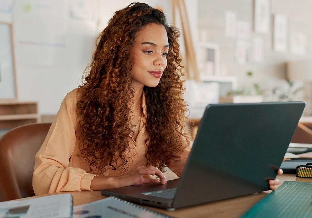 A woman with curly hair sits at a desk, focused on her laptop, surrounded by books and a notepad.