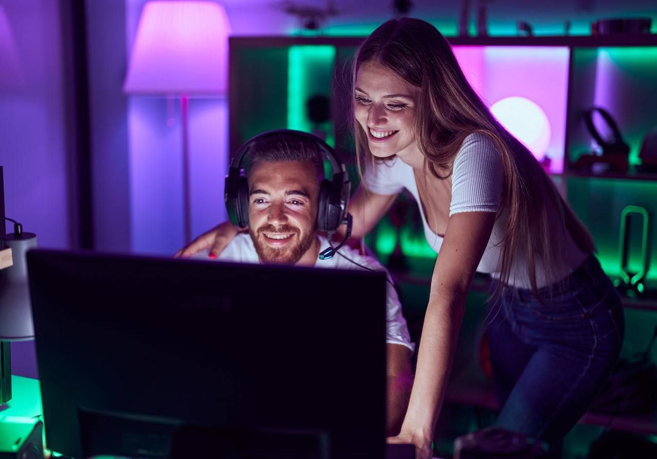 A man with headphones and a smiling woman watch a screen in a colorful gaming room. The ambient glow is purple and green.