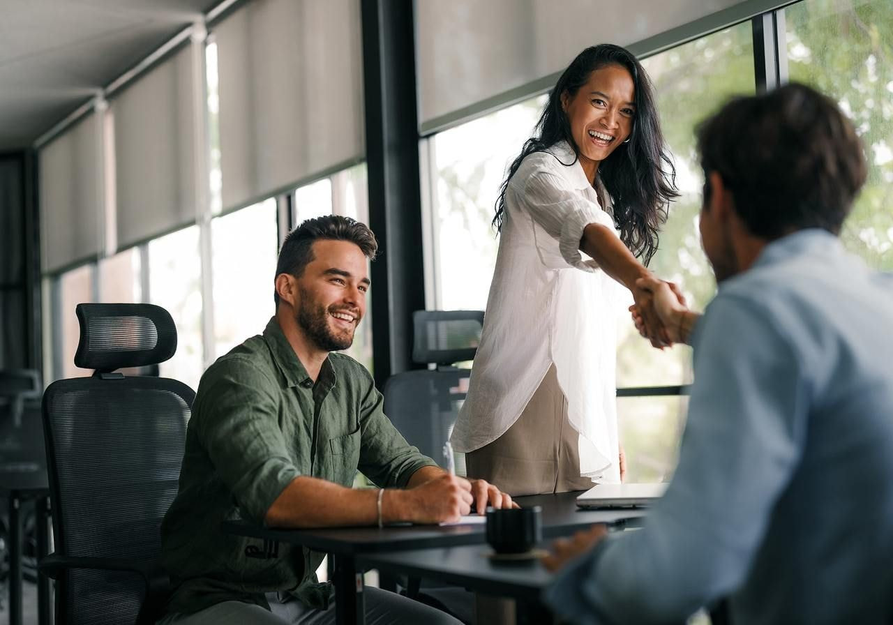 A woman in a white blouse smiles and shakes hands with a man in an office. Another man sits nearby, smiling.