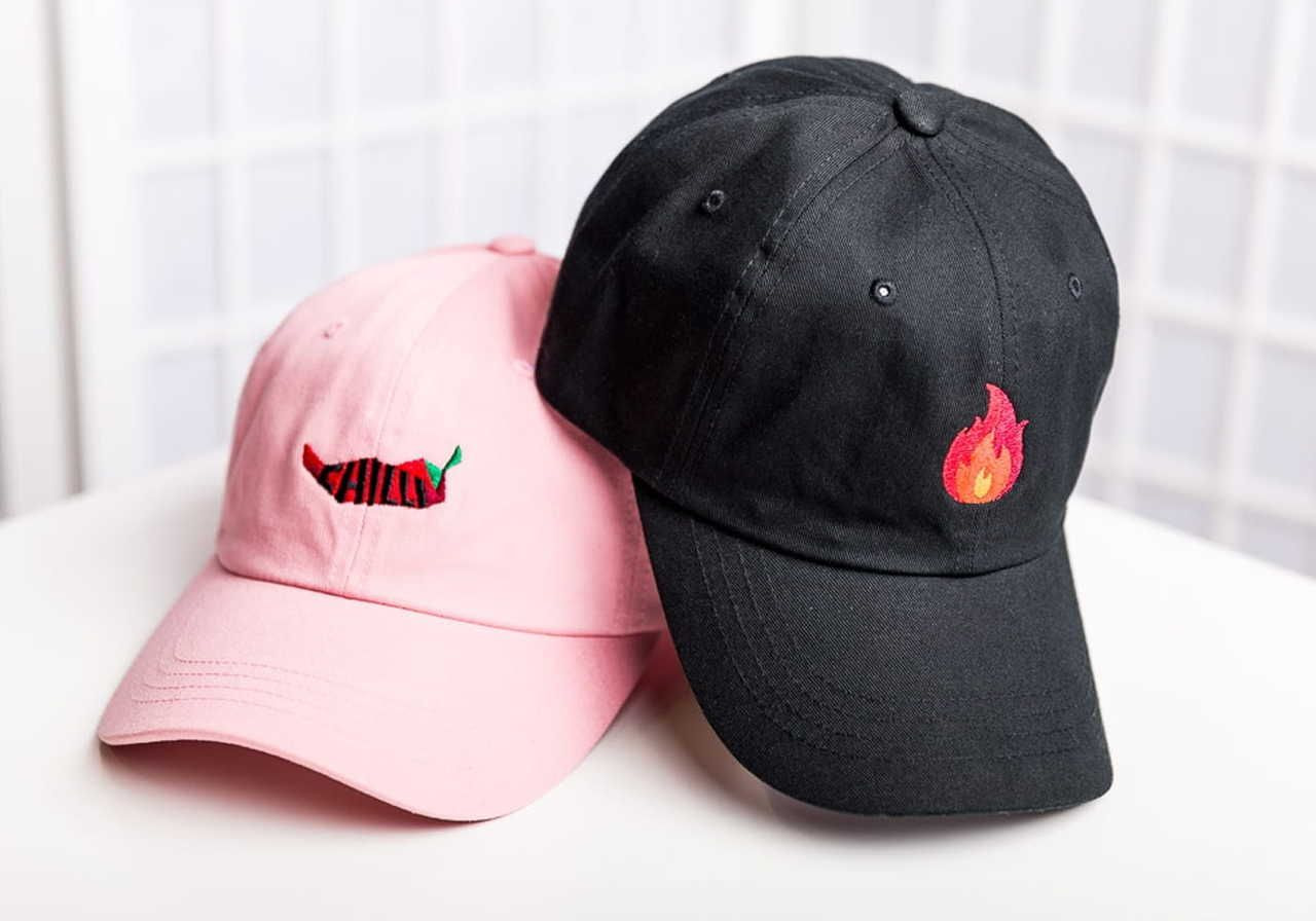 Two baseball caps, one pink with a chili pepper design and one black with a flame design, placed on a table.