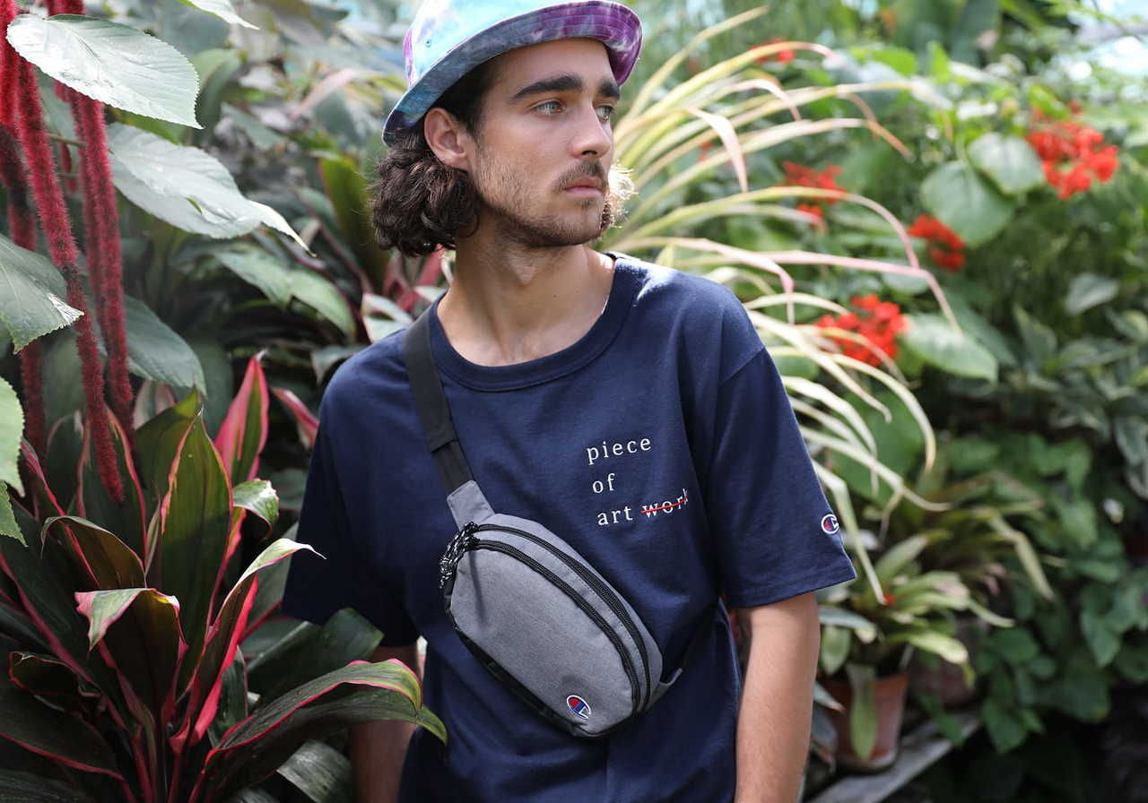 A young man wearing a dark blue t-shirt with text that says "piece of art" and a gray crossbody bag, standing among lush green plants.