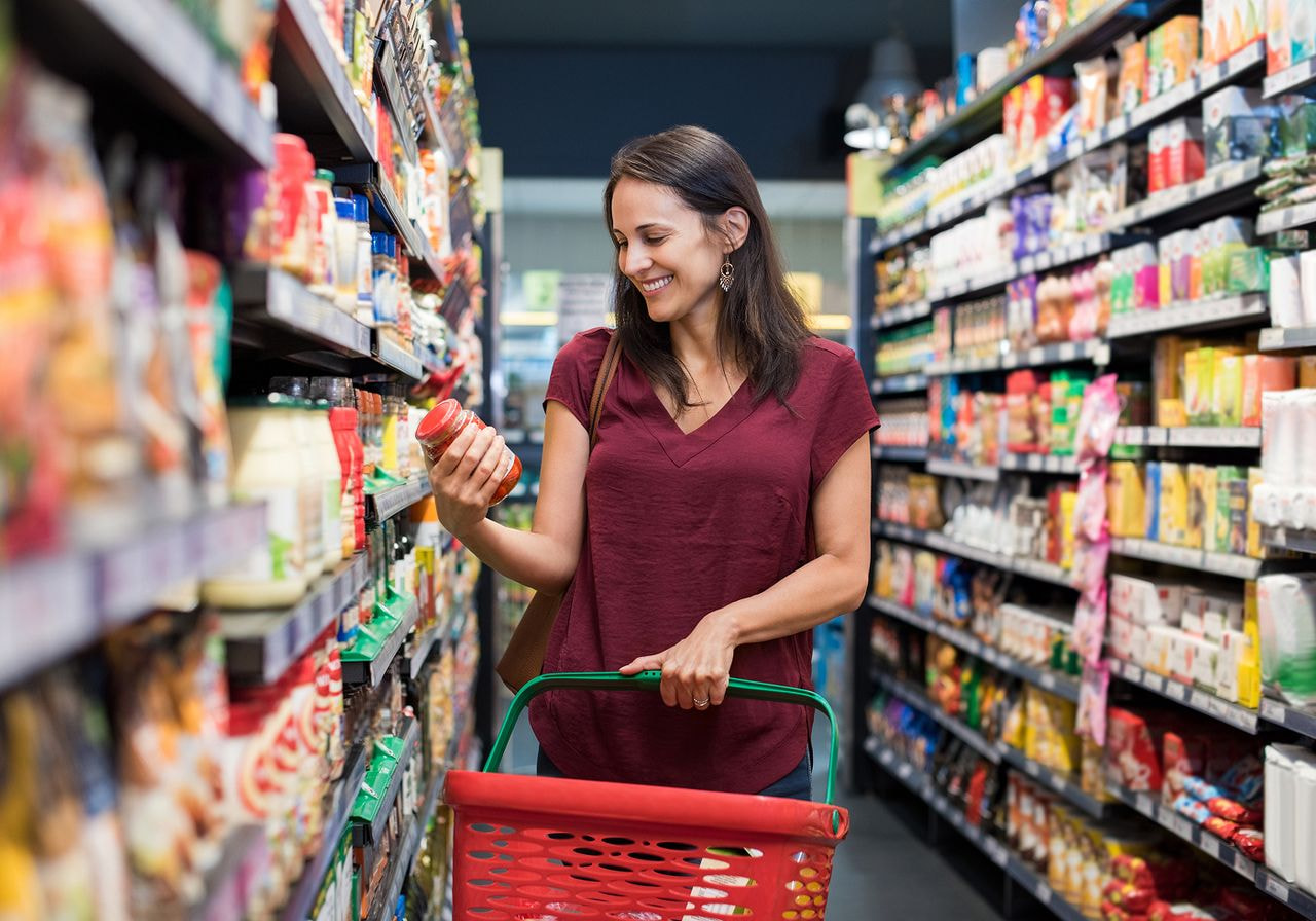 Smiling woman reading a product label while shopping with a basket in a supermarket aisle.