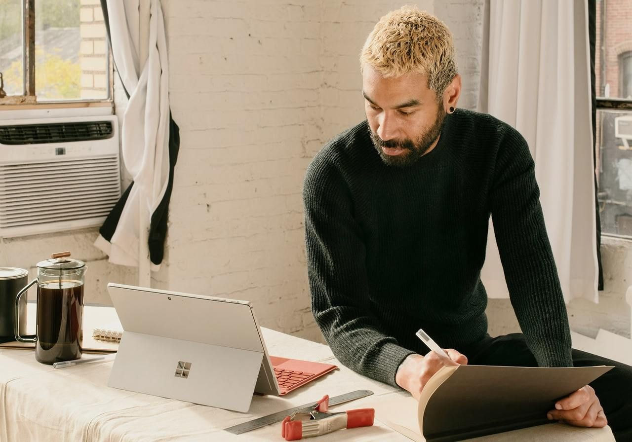 A man is sitting at a desk in a home office and making notes while reading from a laptop “What is merch?”.