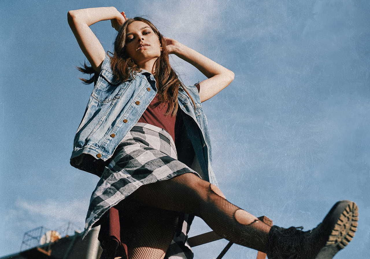 A woman in a denim vest and a checkered skirt poses confidently against a clear blue sky, exemplifying grunge fashion.