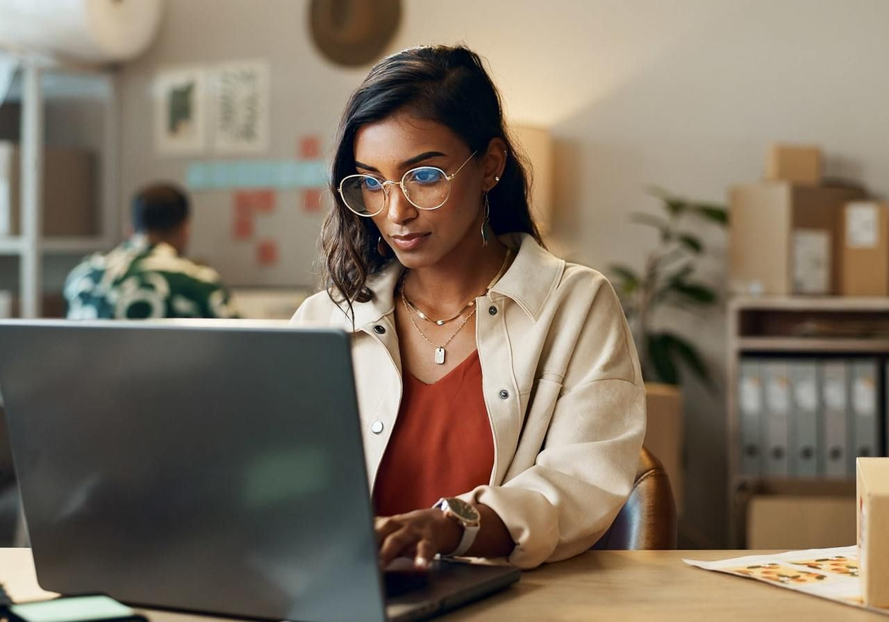 A woman in glasses works on a laptop in an office, researching Black Friday marketing campaigns. She wears a cream jacket over a red top.