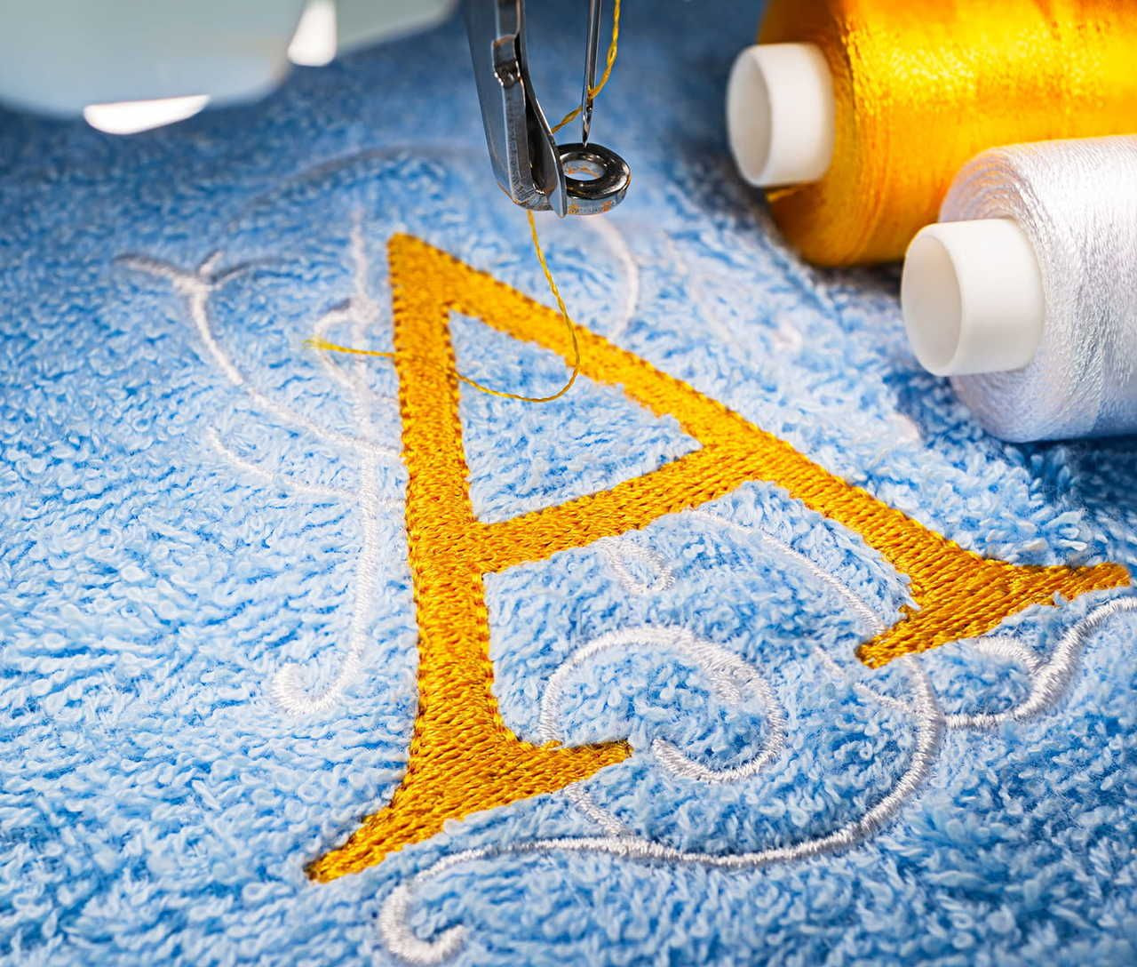 Embroidery machine stitching a yellow letter "A" on blue fabric with decorative white swirls. Nearby, spools of yellow and white thread.