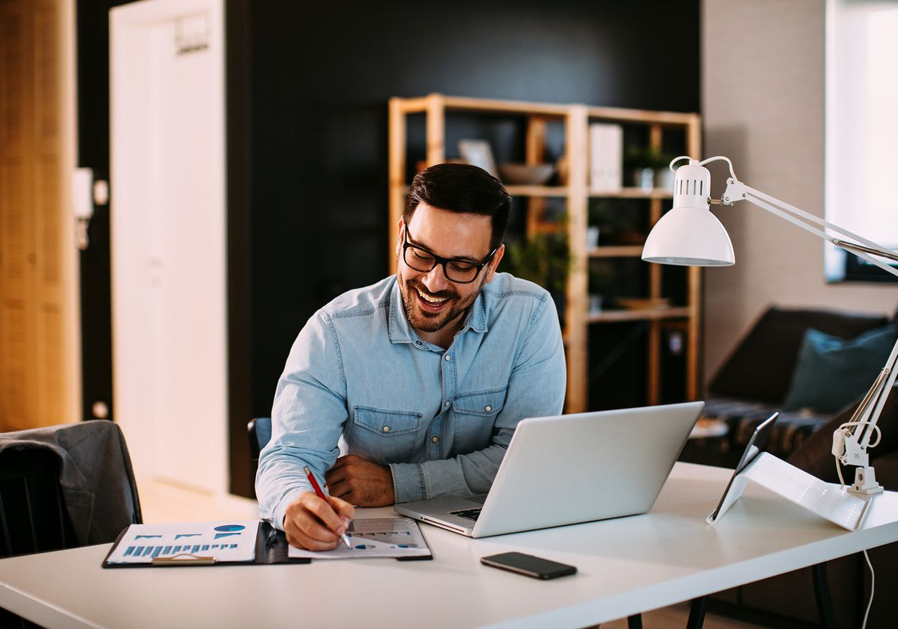 A man sits at a desk, focused on his laptop, with a pen in hand, ready to take notes or write.