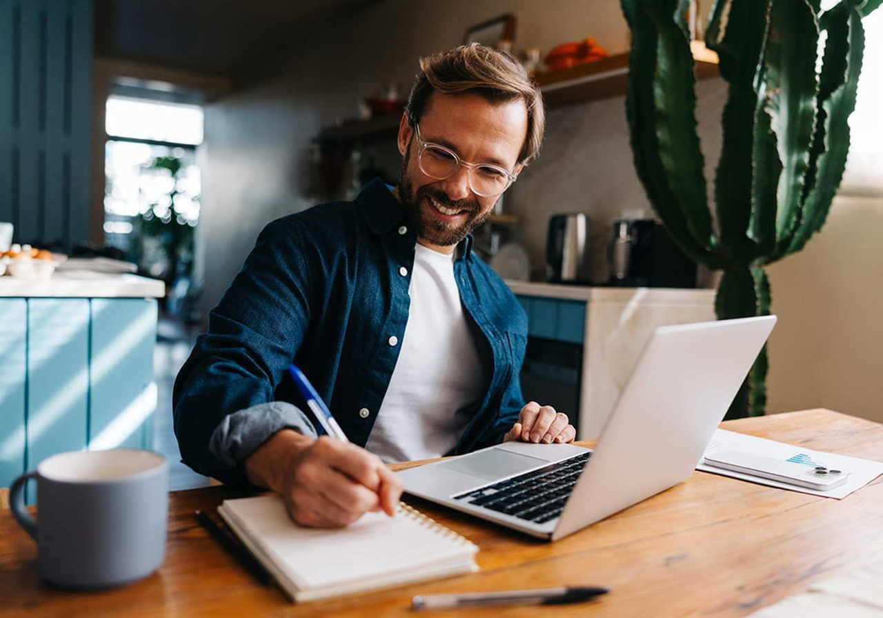 A smiling man with glasses works at a wooden table, writing in a notebook beside a laptop. A large cactus and a mug are nearby.