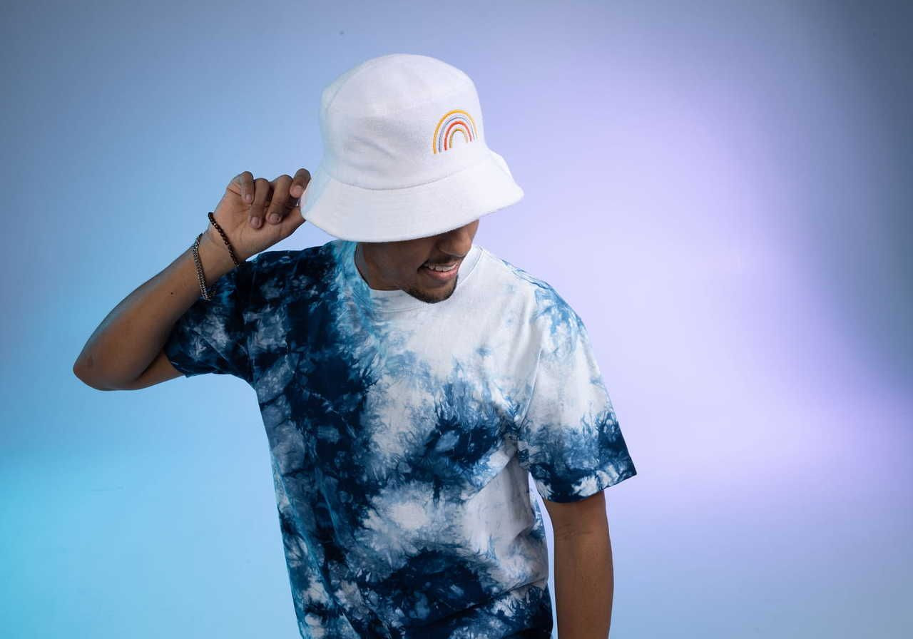A man wearing a white bucket hat with an embroidery rainbow design and a blue tie-dye t-shirt, posing against a gradient background.