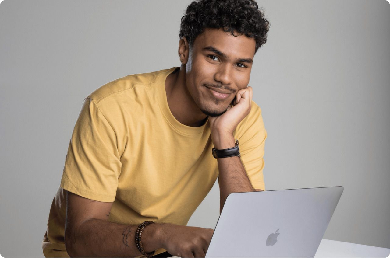 A man in a yellow shirt smiles while working on his laptop.