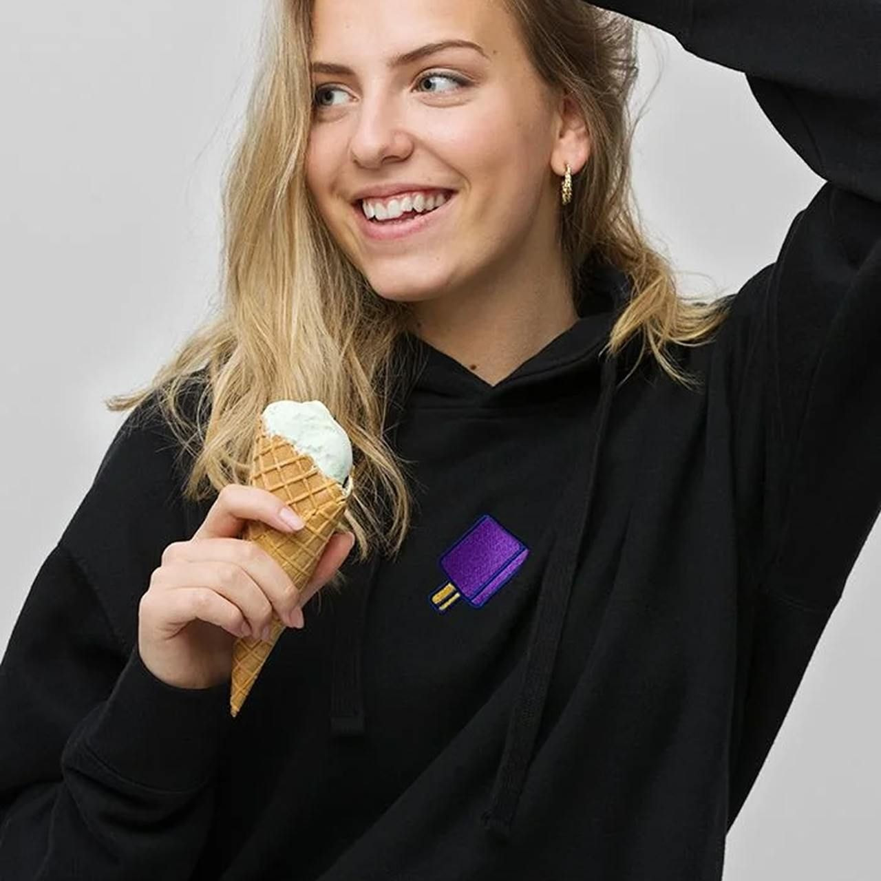 A young woman in a black hoodie with a purple popsicle design smiles while holding an ice cream cone.