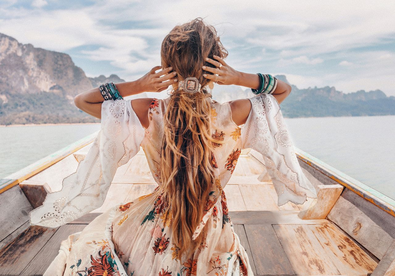 A woman in a floral dress and bracelets sits on a boat, facing scenic mountains and water.