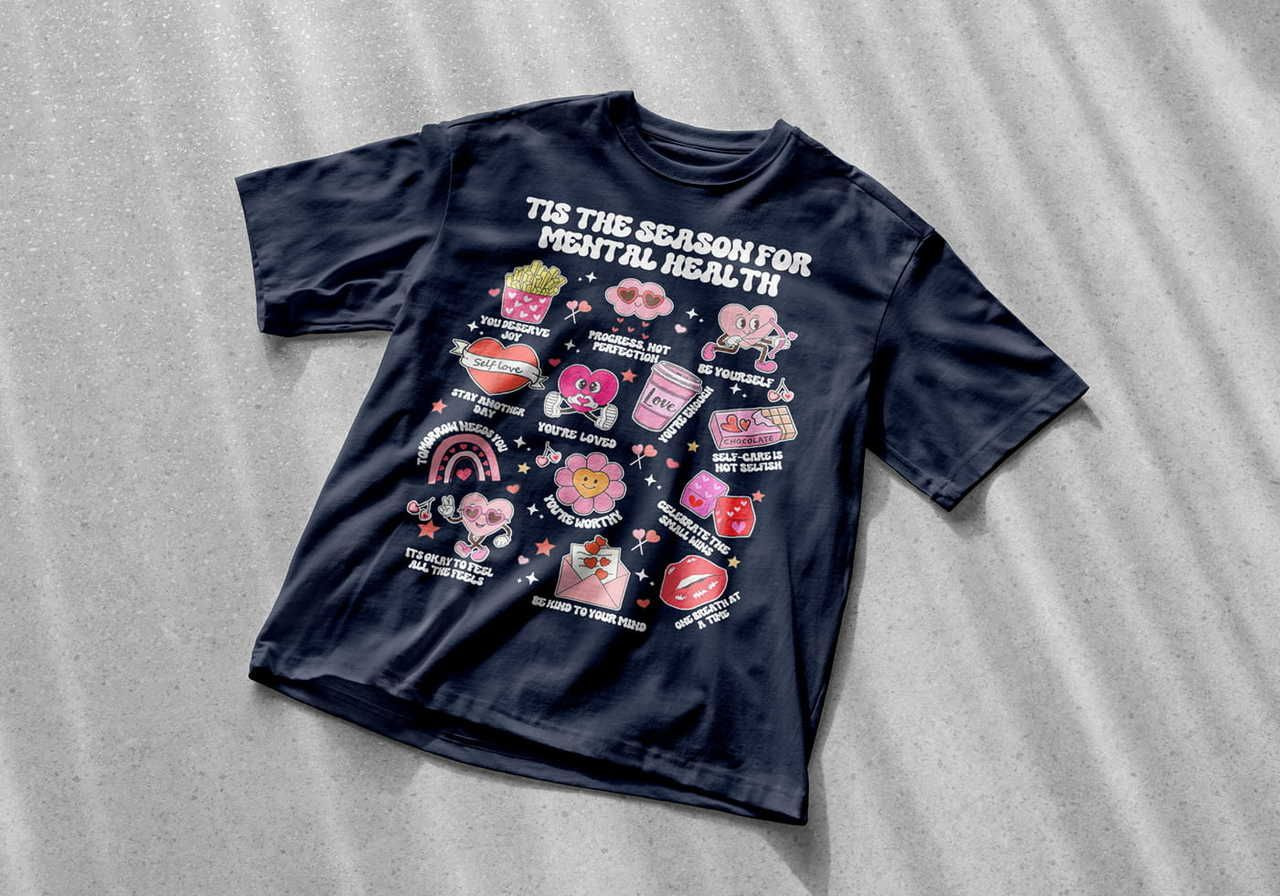 A black t-shirt with colorful cartoon-style illustrations promoting mental health messages like “You’re Loved” and “Be Kind to Your Mind".