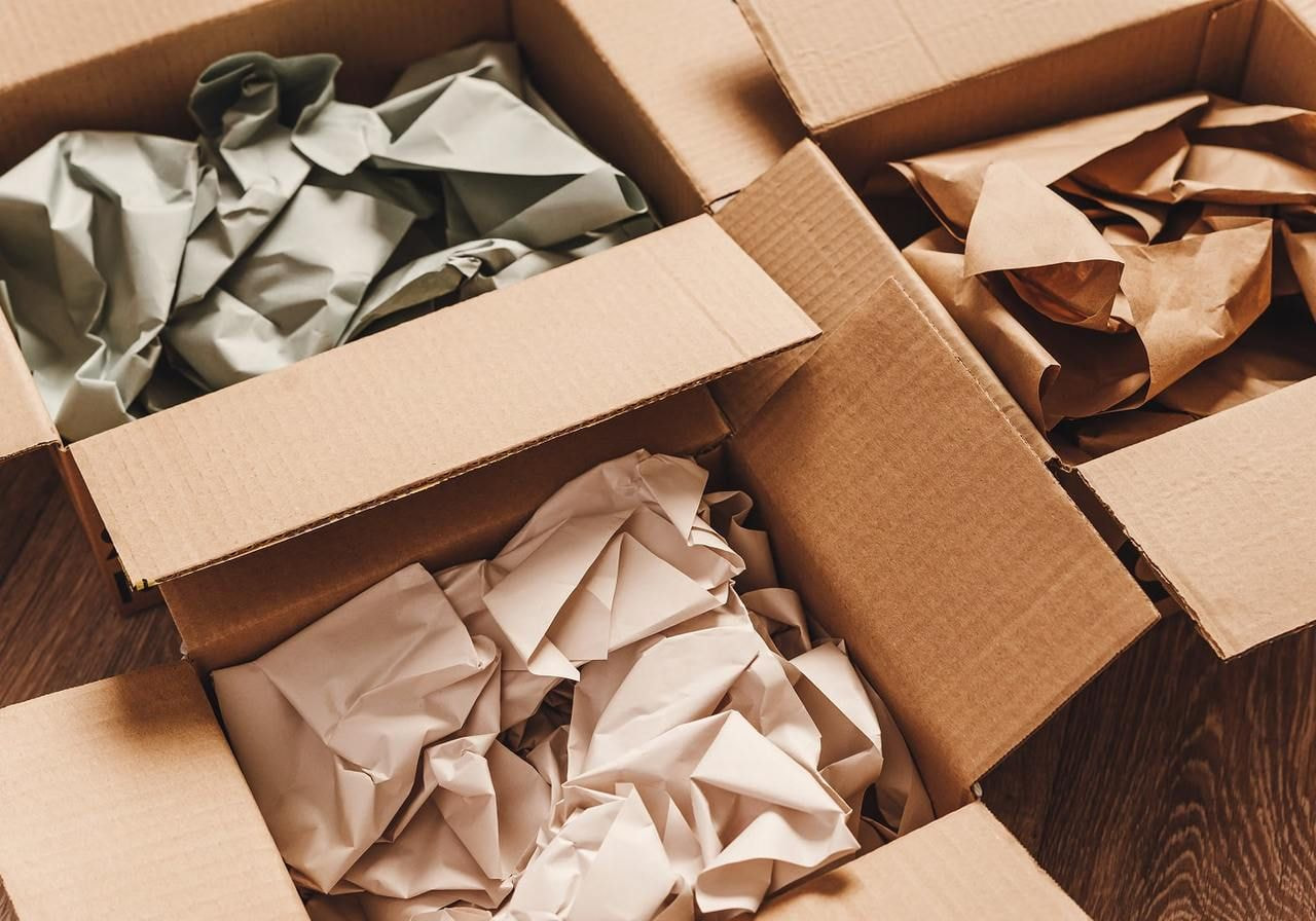 Three open cardboard boxes filled with crumpled packing paper: green, brown, and beige, set on a wooden floor.