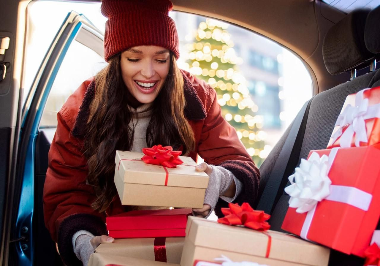 A joyful woman in a red coat and a beanie is placing wrapped gifts in the car, surrounded by festive presents.