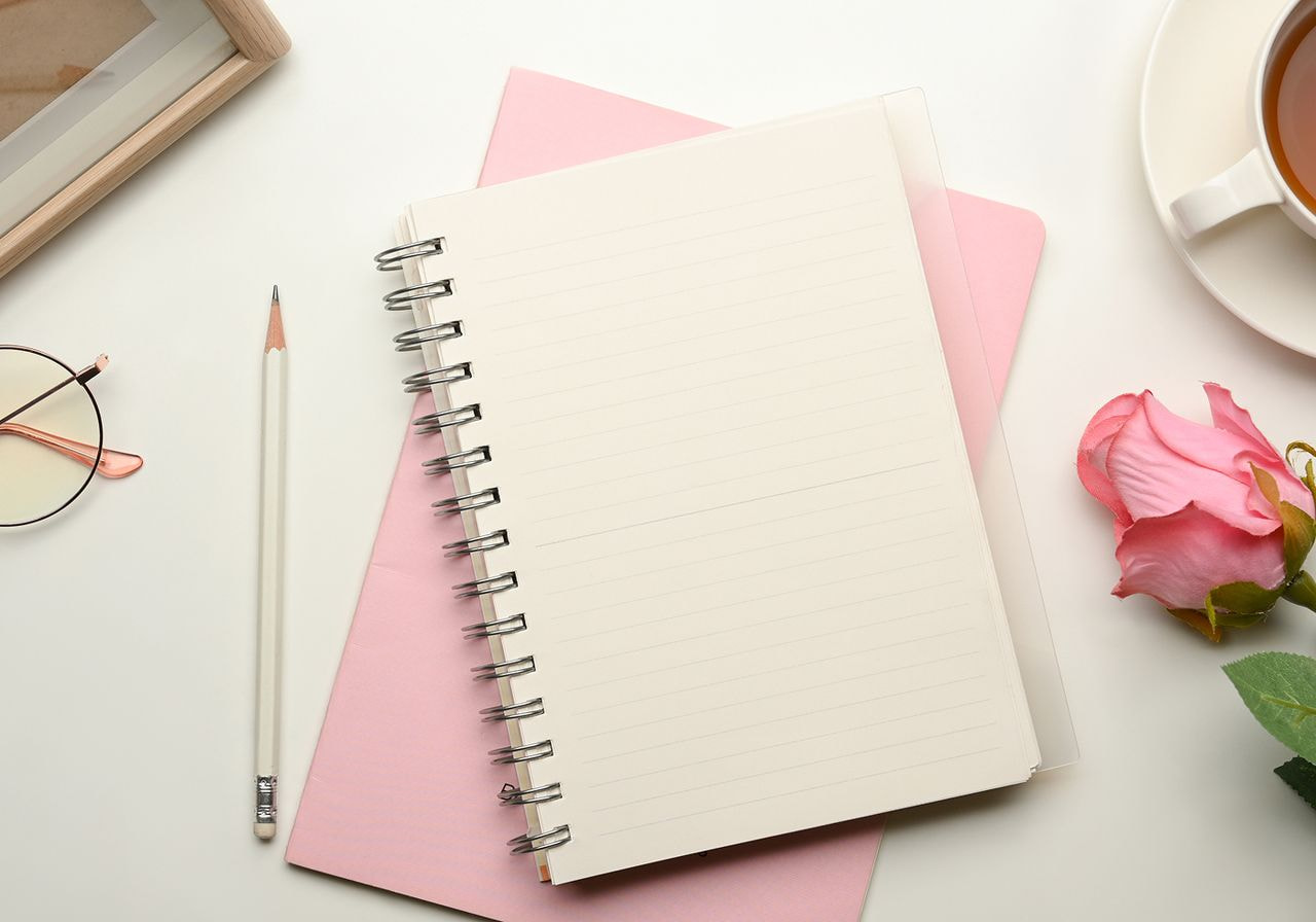A white table displays a notebook, pen, cup of coffee, and a rose, creating a serene workspace setting.